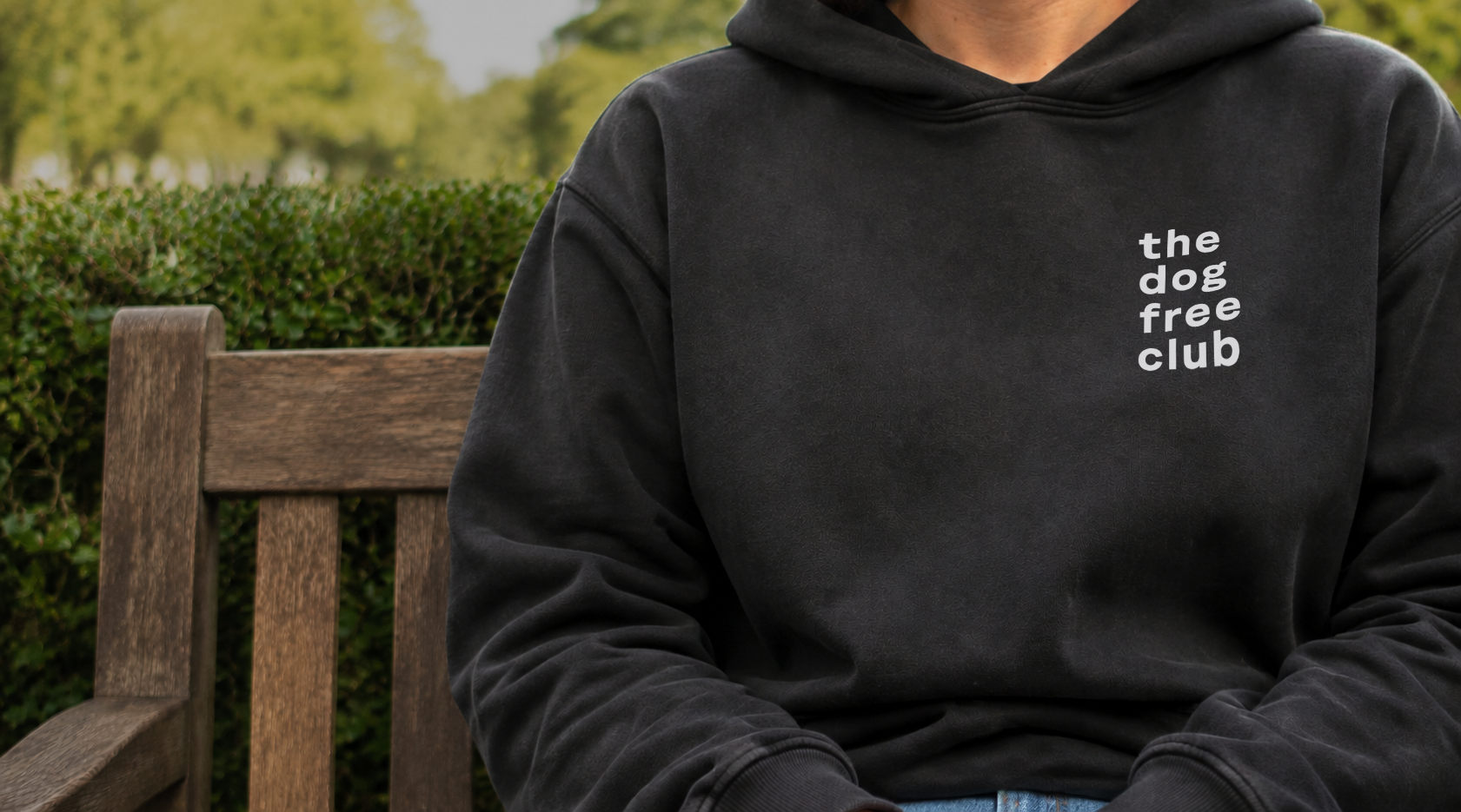 A person sitting on a wooden bench outdoors wearing a faded black hoodie with the text 'the dog free club' printed in white on the left chest. Green hedges and trees are blurred in the background.